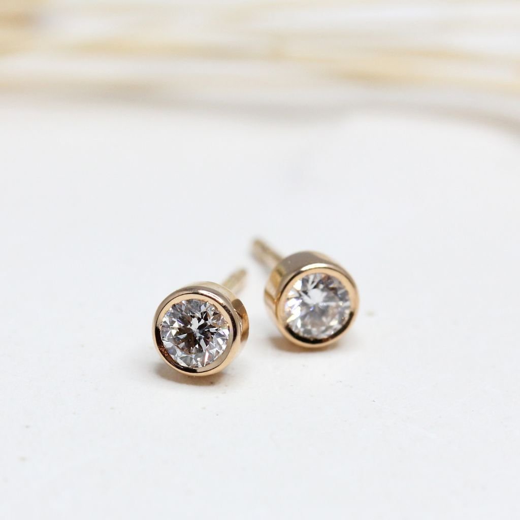 Boucles d'oreille réalisées en or rose-champagne 18 carats Fairmined. Elles sont ornées de sertis clos enserrant deux diamants de synthèse.